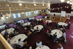 large room with circular tables and people sitting at them listening to a speaker at the front
